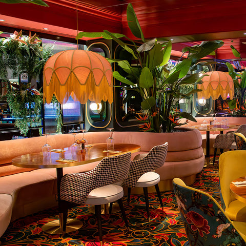 Two large dome shape lampshades in a dusty pink silk with gold trim and long gold fringe hanging in the bar at Grosvenor Casino, a contract setting with a maximalist design and a mix of patterns and floral fabrics as well as plants and flowers