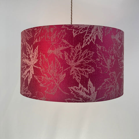 A vibrant fuchsia red drum lampshade with a delicate maple leaf pattern in a rich jacquard fabric. The shade features a frosted clear inner lining and a sleek beach gold frame, adding a touch of modern elegance to any room. Available in various sizes, it is designed to complement both contemporary and traditional interiors