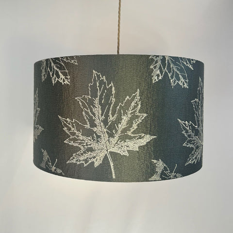 A sophisticated green drum lampshade featuring intricate maple leaf embroidery in warm, fall colours. The rich dark green fabric is paired with a frosted clear inner lining and a stylish beach gold frame. This elegant lampshade is designed to bring a luxurious touch to any space
