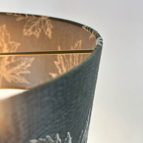 A close-up of a sophisticated green drum lampshade featuring intricate maple leaf embroidery in rich dark green fabric is paired with a frosted clear inner lining and a stylish beach gold frame. This lampshade is designed to bring a luxurious elegance to any space.