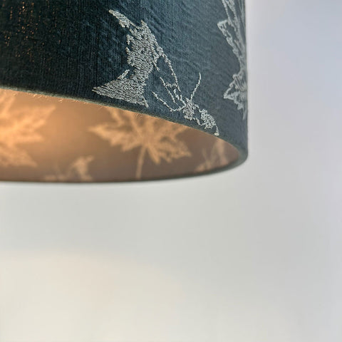 A close-up of a luxury fabric with intricate maple leaf embroidery in warm, fall colours on a lampshade. The rich dark green fabric is paired with a frosted clear inner lining. This sophisticated lampshade is designed to bring an elegant touch to any space.