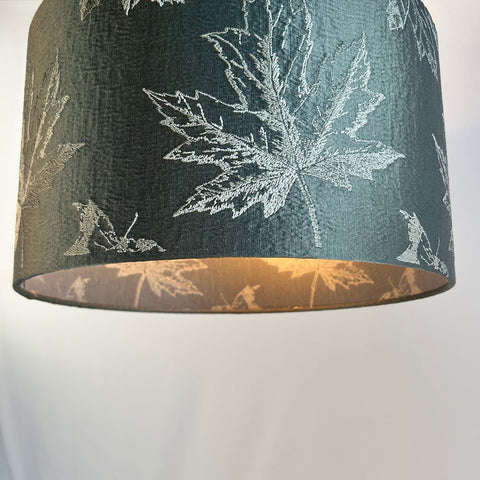 A close-up of a sophisticated green drum lampshade featuring intricate maple leaf embroidery in warm, fall colours. The rich dark green fabric is paired with a frosted clear inner lining showing off the pattern inside and out. This luxury lampshade is designed to bring a touch of elegance to any space.