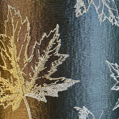 A close-up of a sophisticated green fabric featuring intricate maple leaf embroidery in warm, fall colours. The rich dark green with silver fabric shines on an elegant lampshade and is designed to bring a luxurious touch to any space.