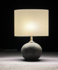 A table lamp with a stoneware base and a silk drum lampshade, illuminated, set against a dark background.