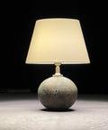 A table lamp with a stoneware base and a silk empire lampshade, illuminated, set against a dark background.
