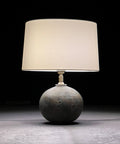 A table lamp with a stoneware base and a silk French drum lampshade, illuminated, set against a dark background.