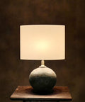 A table lamp with a stoneware base and a silk drum lampshade, illuminated, set against a dark background.