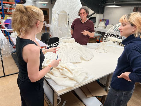 Three people discussing around  a large white lampshade on a work table in a workshop setting.